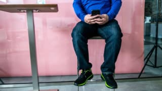 A person in jeans sits on a stool and is looking at their phone in front of a pink wall