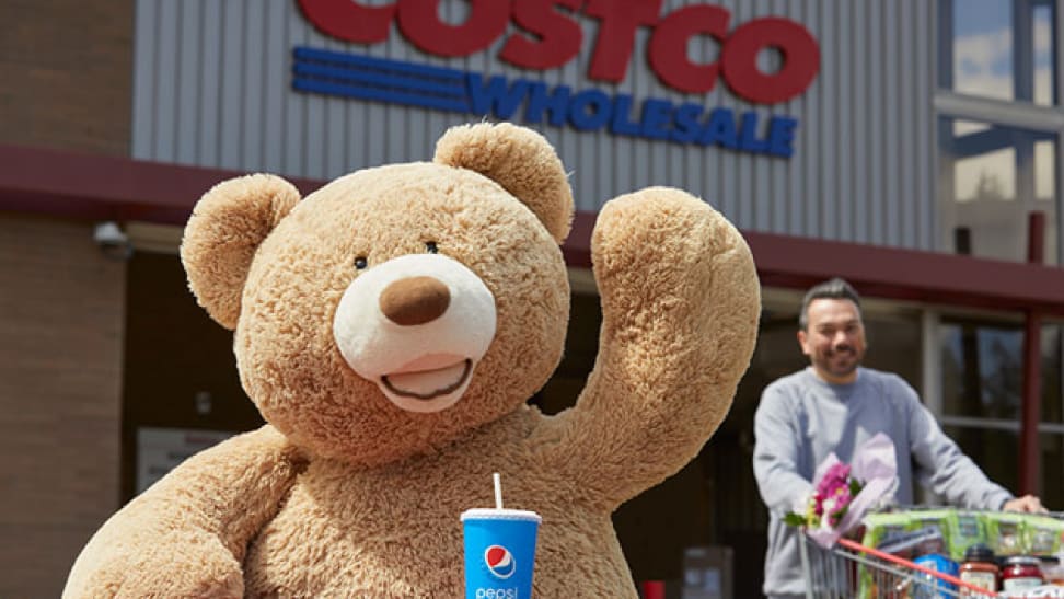 A large teddy bear is standing in front of a Costco while a shopper pushes a cart nearby. The bear has a pepsi cup.