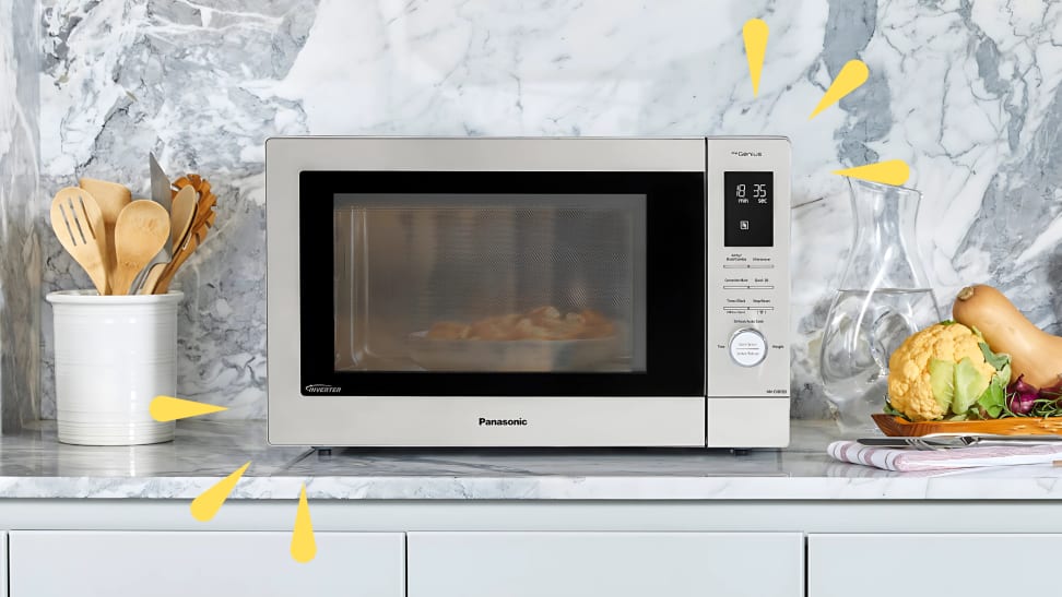 A Panasonic microwave sits on a marble kitchen counter surrounded by cooking utensils and fresh vegetables.