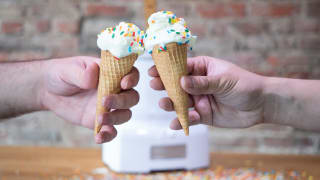 Two hands holding ice cream cones with vanilla ice cream and rainbow sprinkles cheers in front of an ice cream maker on a wood counter in front of an exposed brick wall.