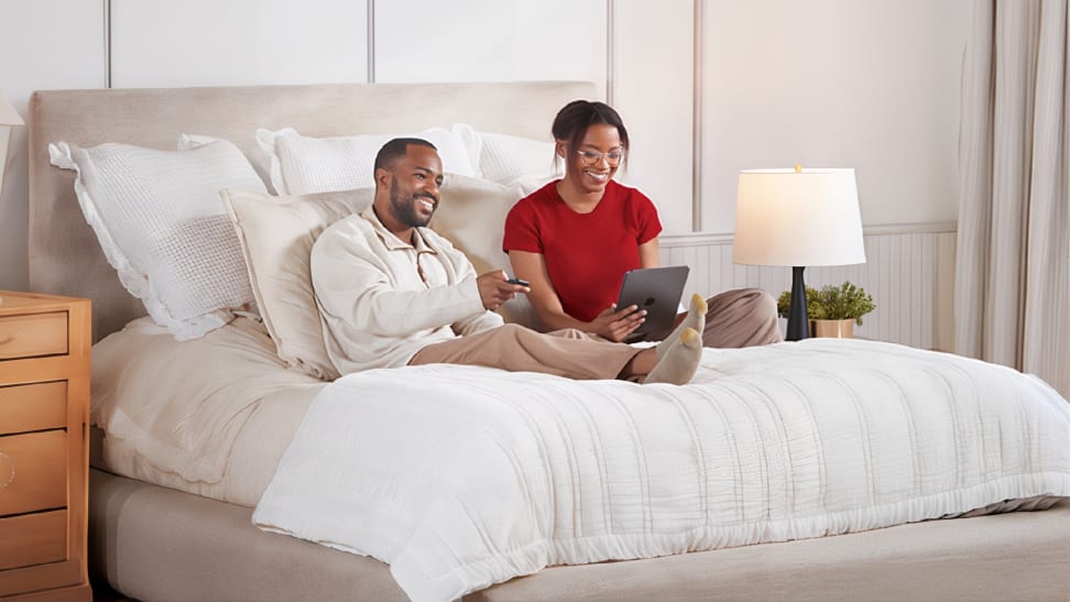 A smiling couple relaxes on a neatly made bed with cream-colored bedding, as one person holds a remote and the other uses a tablet. A wooden nightstand with a lamp and potted plant sits beside the bed in a bright, cozy bedroom.